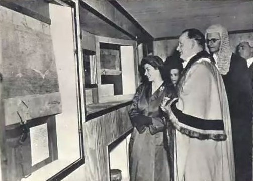 Historical image of a Princess Elizabeth in a formal outfit, accompanied by men in robes, observing displayed artifacts in a museum setting.