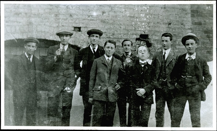 A historical black and white photograph of nine young men dressed in early 20th-century attire, standing together in front of a brick wall.