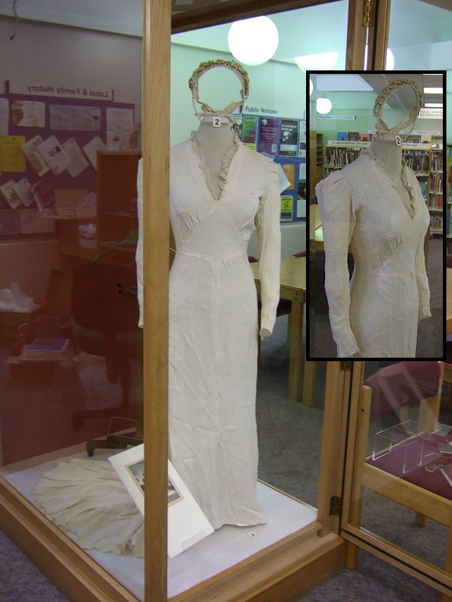 A vintage wedding dress on display in a museum exhibit, featuring intricate fabric details and a long train, with a close-up view in an inset image.