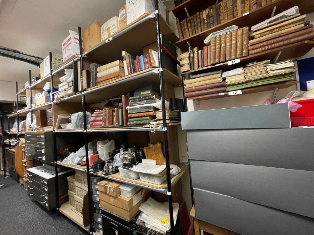 A view of the Archive Store, featuring shelves filled with various books, boxes, and artifacts covered in protective materials.