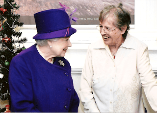 The Queen and Hester Davenport smiling and engaging in conversation, one wearing a purple hat and coat, the other in a light-colored blouse, with a Christmas tree in the background.