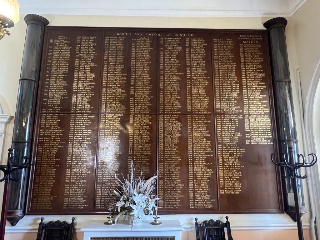 A wooden wall display in the Guildhall featuring the names of past Bailiffs and Mayors of Windsor, with gold lettering against a dark background. A decorative floral arrangement is placed in the foreground.