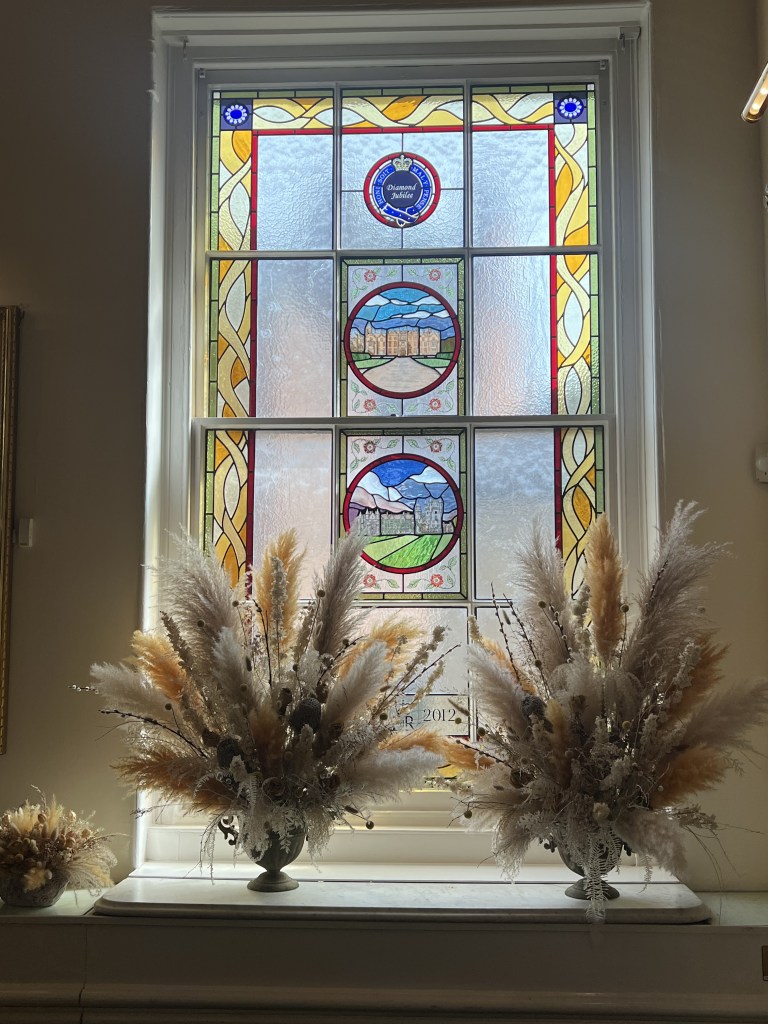 Stained glass window featuring royal symbols and landscape imagery, accompanied by decorative floral arrangements in vases.