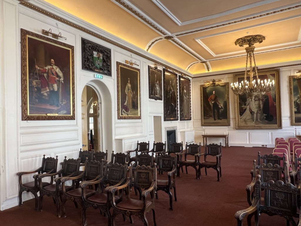 Interior of the Windsor Guildhall featuring ornate portraits on the walls, a chandelier, and wooden seating arranged for an event.
