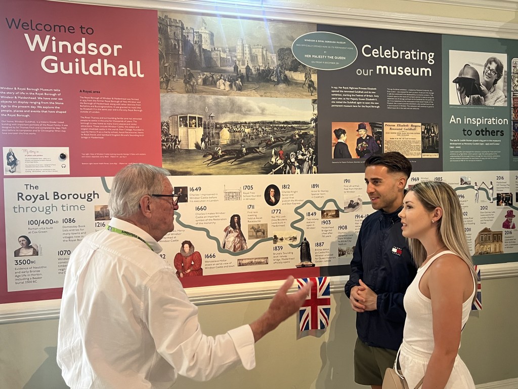 A volunteer with two visitors engaged in conversation in front of an exhibition wall at Windsor Guildhall, displaying historical information and images related to the Royal Borough.