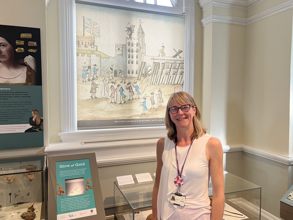 Stephanie, a museum staff member stands in front of a historical display featuring a tapestry and artifacts at the Windsor & Royal Borough Museum.