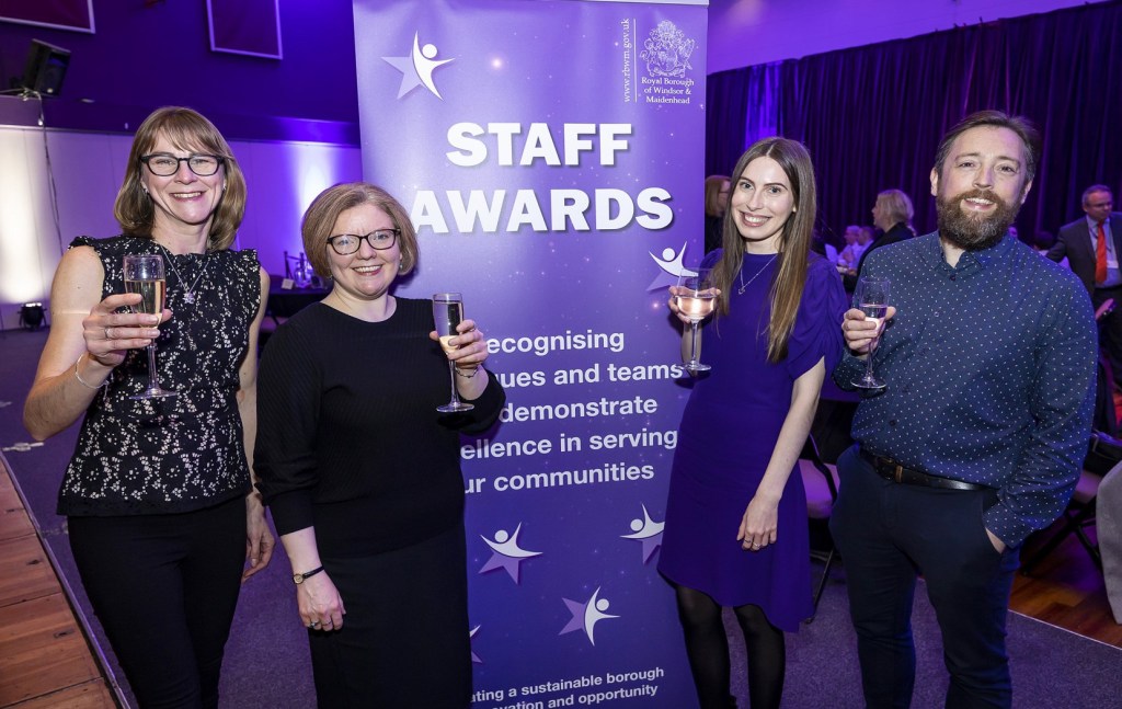 Four staff members celebrating during the Staff Awards event, holding drinks and smiling in front of a banner highlighting staff excellence.