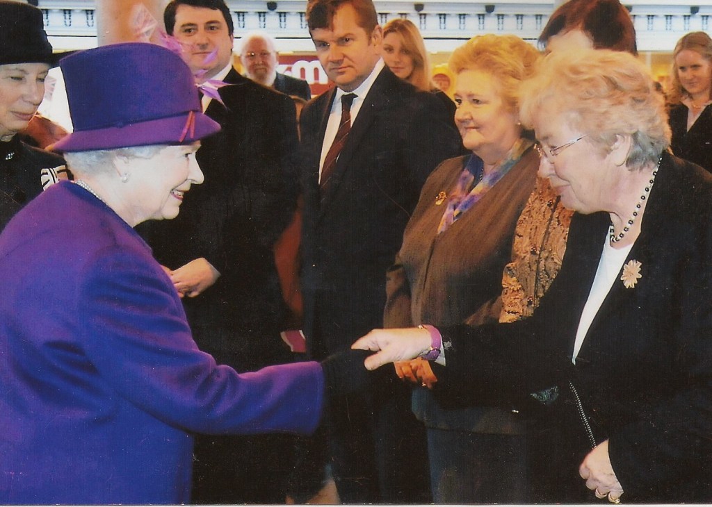 Her Majesty Queen Elizabeth II meeting Brigitte Mitchelll at a public event, with others observing in the background.