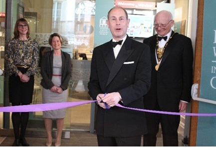 A formal event featuring Prince Edward in a tuxedo cutting a purple ribbon at a museum opening, accompanied by guests and museum staff in professional attire.