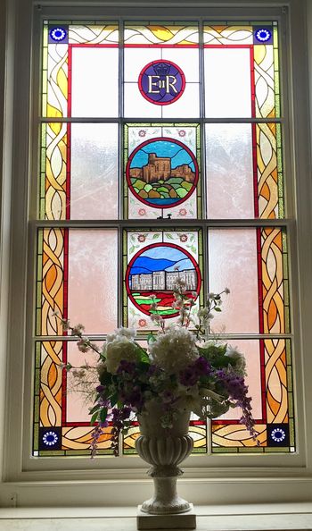 A stained glass window featuring decorative elements and royal symbols, alongside a floral arrangement in a vase.