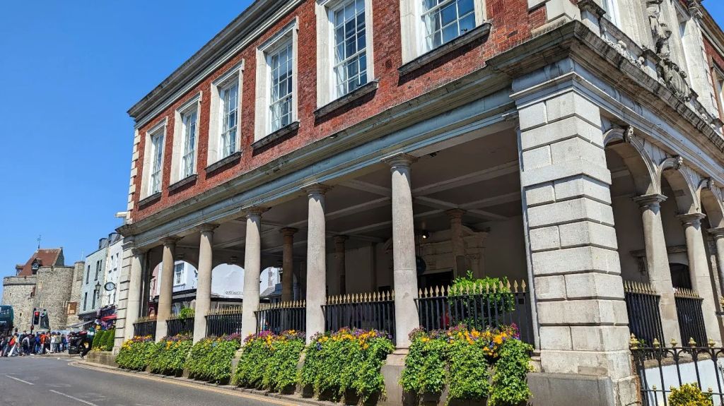 View of the Windsor Guildhall, a historic building with a combination of red brick and stone architecture, featuring tall columns and flower beds in the foreground.