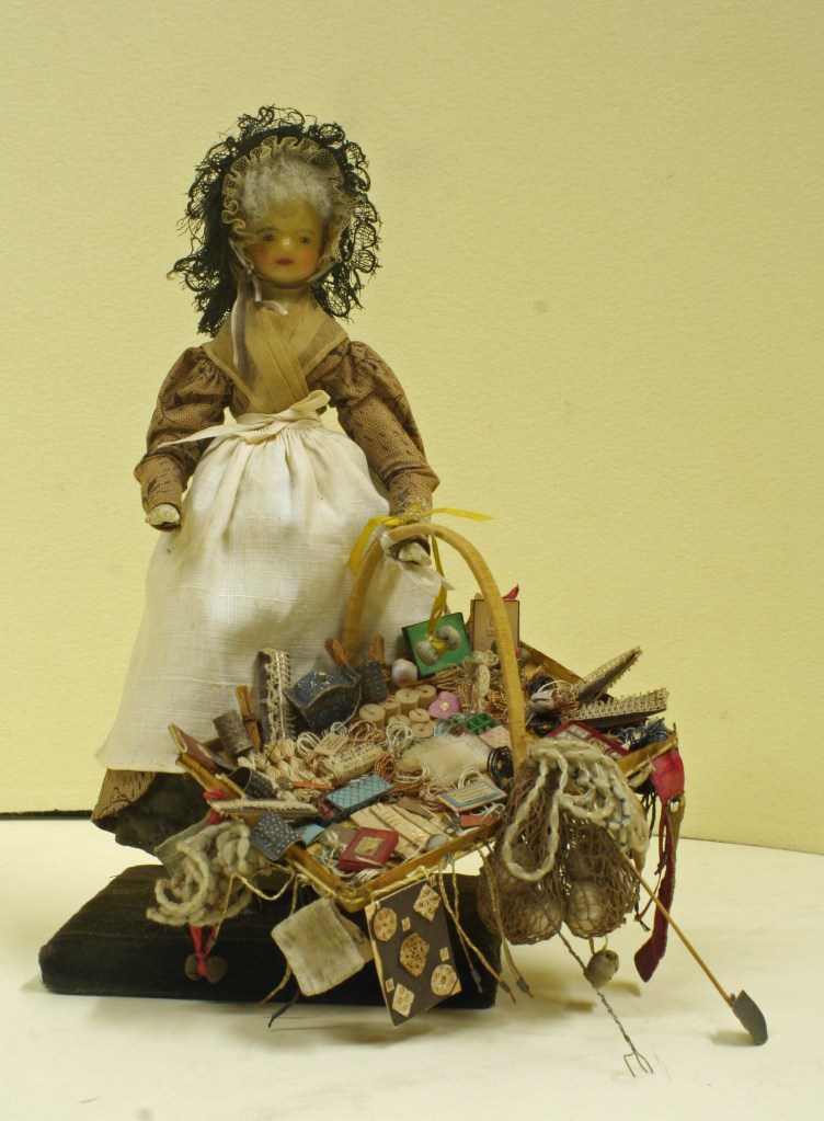 A vintage Georgian pedlar doll displaying a basket filled with various small items and trinkets, set against a light yellow background.