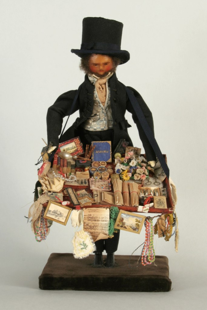 A Georgian Pedlar Doll dressed in traditional attire, holding a display of various miniature goods and trinkets.