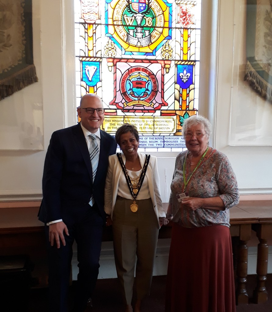 Three people stand in front of a stained glass window, celebrating the Festival of Britain. The individuals are smiling, dressed in formal attire, with one wearing a mayoral chain. The window features colorful emblems and designs, commemorating a historical event.