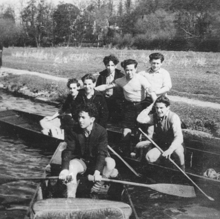 Group of young Holocaust survivors enjoying a day on the river, sitting in a rowing boat and posing for the camera, with trees and grass in the background.
