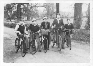 A black and white photograph of five children standing on a dirt road, each with a bicycle. They are dressed in vintage clothing, with one boy wearing a straw hat, and trees and a fence are visible in the background.