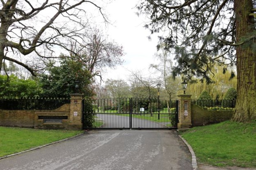 Entrance to Woodcote Place, the former site of Woodcote House, featuring black gates surrounded by trees and green grass.