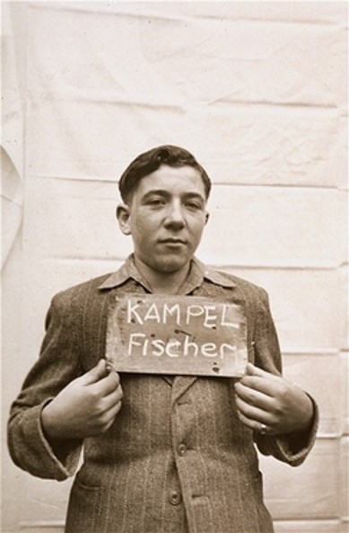 Photograph of a young boy holding a sign that reads 'KAMPEL Fischer', wearing a simple, wartime jacket against a plain backdrop.