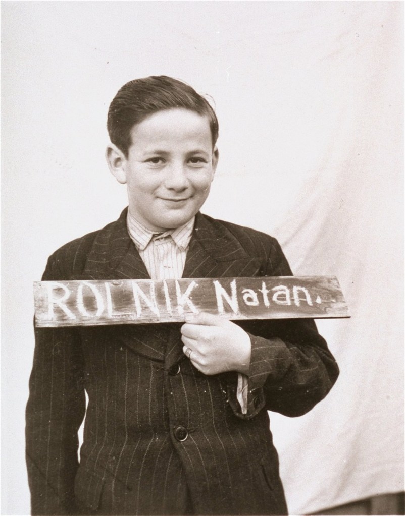 A young boy named Natan Rolnik, wearing a suit and holding a sign with his name, poses for a photograph. The background is a plain white wall.