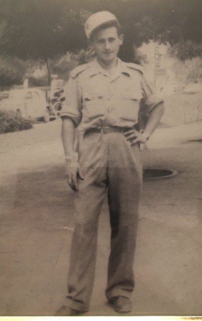 Sam Freiman as a young man dressed in military uniform stands confidently on a street, with trees and cars in the background.