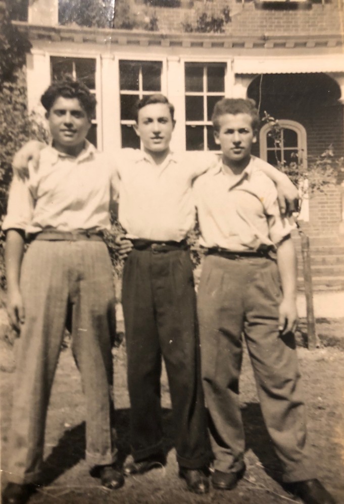 Three young men standing together outdoors, wearing casual clothing, with a building in the background.