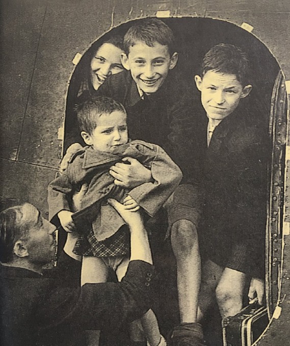 A historical black and white photograph showing a group of four children, including a young girl and a toddler, being assisted by an adult in what appears to be an airplane entrance, symbolizing their journey as Holocaust survivors.