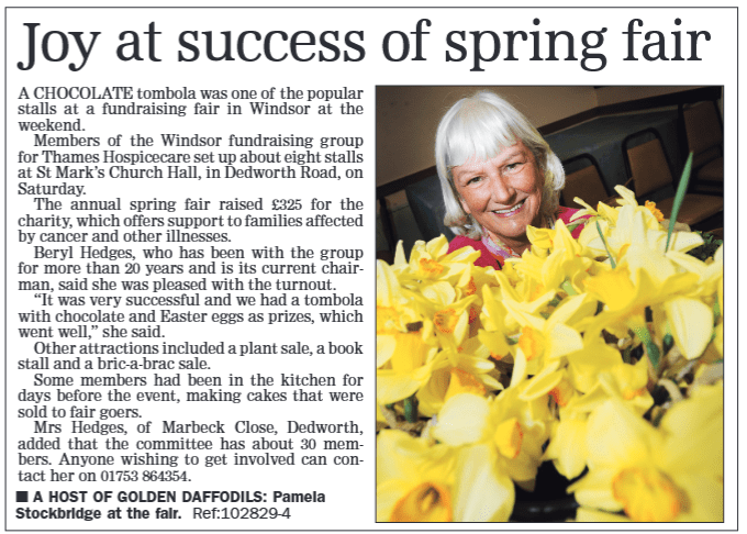 A smiling woman with light hair is holding a bunch of yellow daffodils at a fundraising fair. The background features stalls and people attending the event.