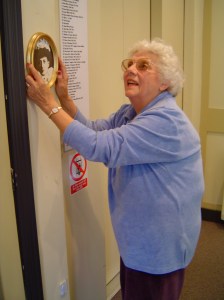 Beryl Hedges, with curly white hair, is hanging a framed picture on a wall. She is wearing glasses and a blue cardigan, smiling as she adjusts the picture.