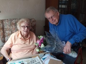 Beryl Hedges sitting in a chair, smiling and holding a bouquet of flowers, with Len  beside her in a blue shirt.