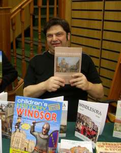 Elias Kupfermann smiling, holding one of his books, sitting at a table with various history publications and a wooden background.