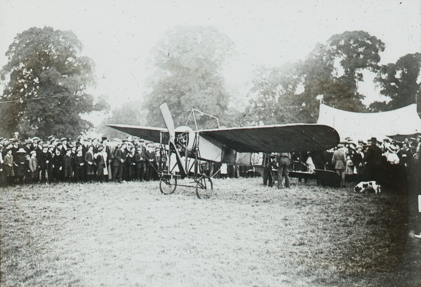 A historic black-and-white photograph showing a crowd gathered around an early biplane on an open field, with trees and a tent in the background.