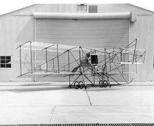 A historical biplane aircraft displayed in front of a hangar, showcasing its frame and structure.