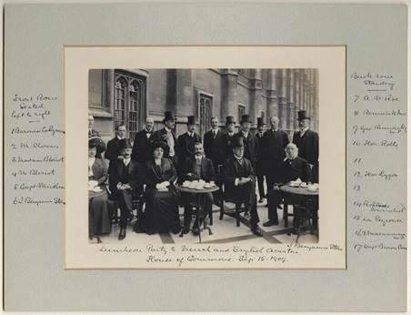 A historical black-and-white photograph of a large group of elegantly dressed men and women seated and standing around tables with tea and refreshments, taken at a formal gathering in front of an old building.