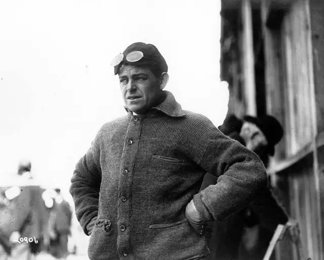 Black and white image of a man wearing goggles and a thick sweater, standing with his hands on his hips, appearing pensive.