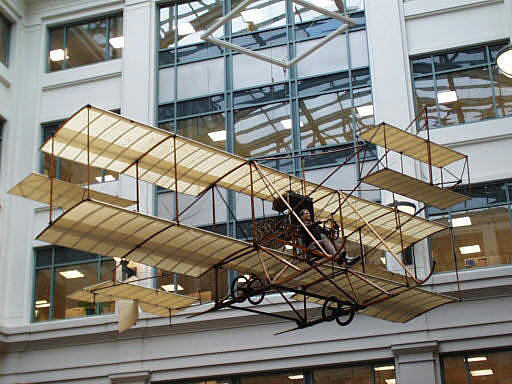 A historic biplane displayed indoors, showcasing its wooden frame and fabric wings in front of large windows.
