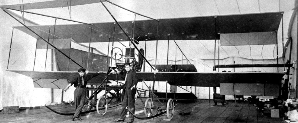 Two men standing beside a biplane inside a hangar, working on its structure.