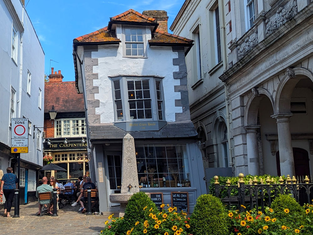The Crooked House, a leaning timber-framed building in Windsor, with outdoor seating at a nearby pub and colorful flowers in the foreground.