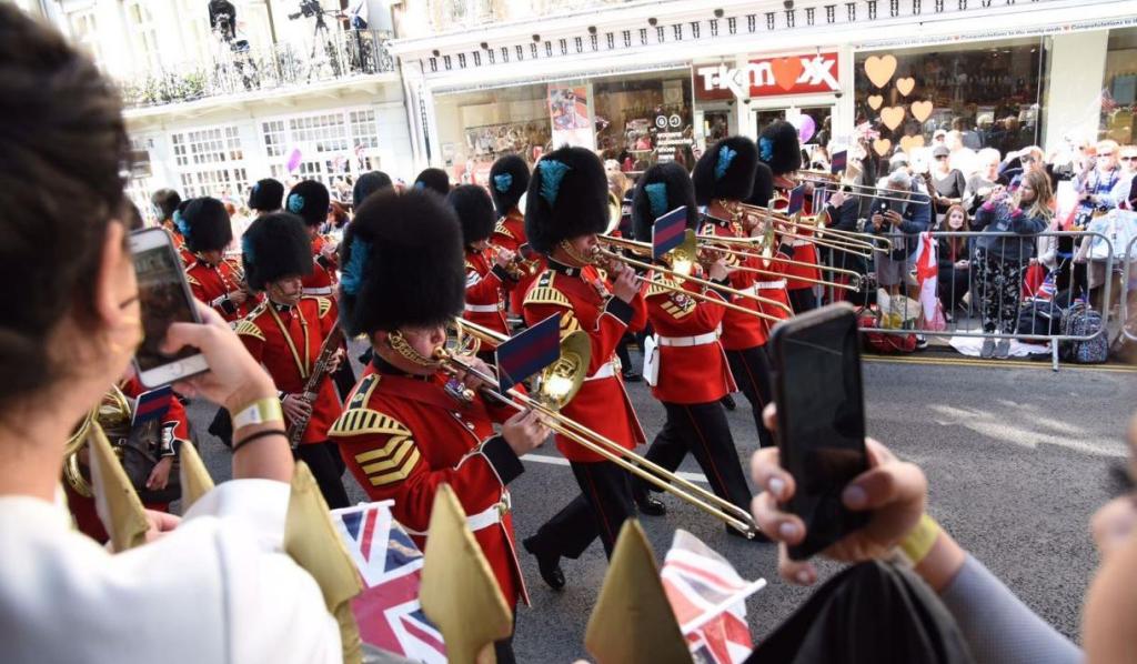 A group of British soldiers in red uniforms and bearskin hats perform during a ceremonial parade, with spectators watching and taking photos in the background.