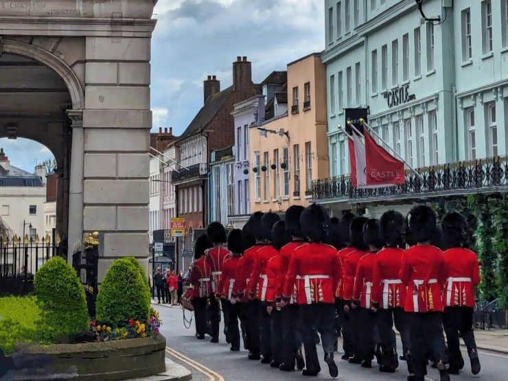A group of British soldiers in traditional red uniforms and black bearskin hats marching passed tge Guildhall in Windsor, surrounded by buildings and greenery.