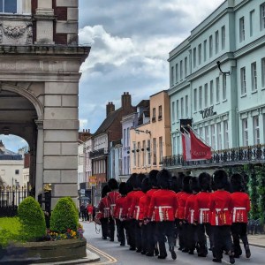 A group of soldiers in red uniforms and bearskin hats marching down a street lined with historic buildings in Windsor.