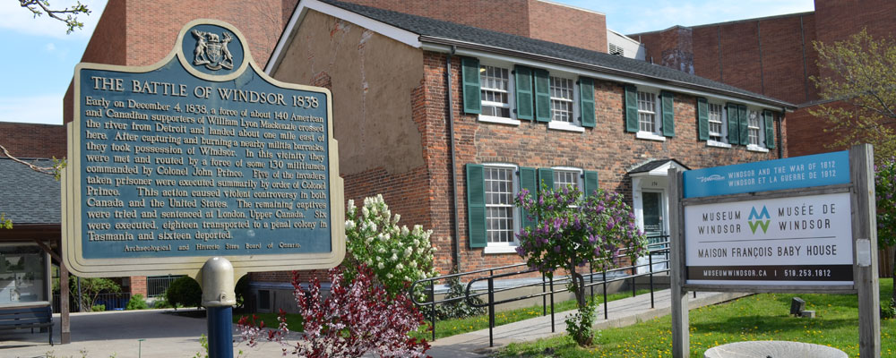 Signage detailing the Battle of Windsor 1838 in front of the Museum Windsor and the Maison François Baby House, showcasing historical significance and architecture.