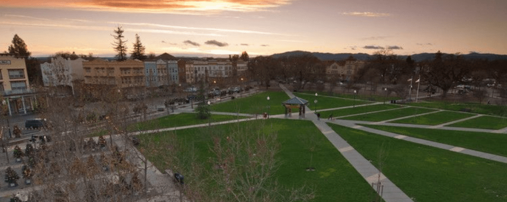 A panoramic view of Windsor Town Green in California at sunset, featuring open green spaces, pathways, and surrounding buildings.