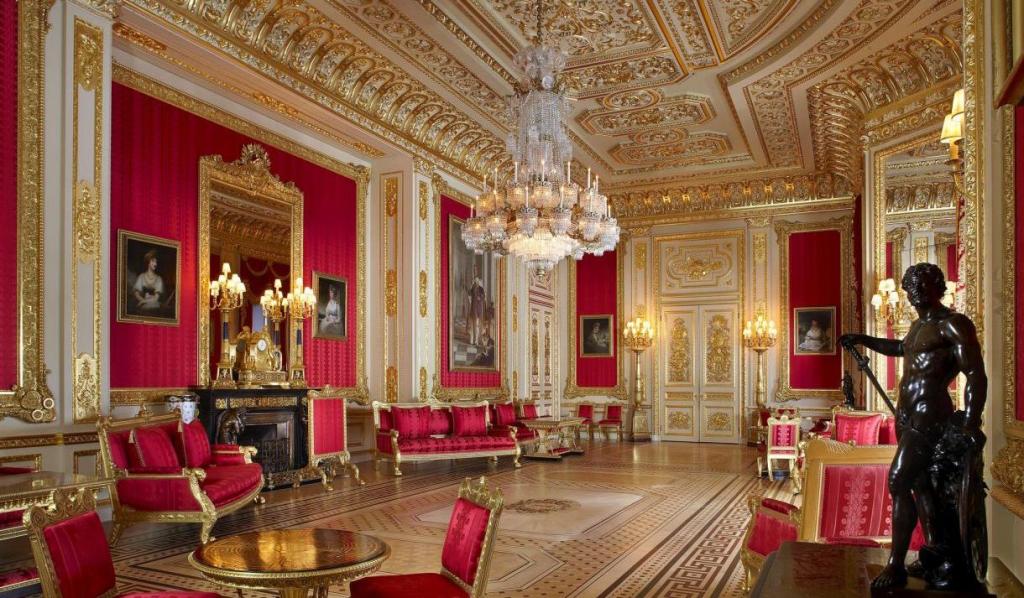 An opulent room featuring ornate gold decorations, red walls, a grand chandelier, and luxurious seating. The room also includes paintings and decorative artworks, creating a regal atmosphere.