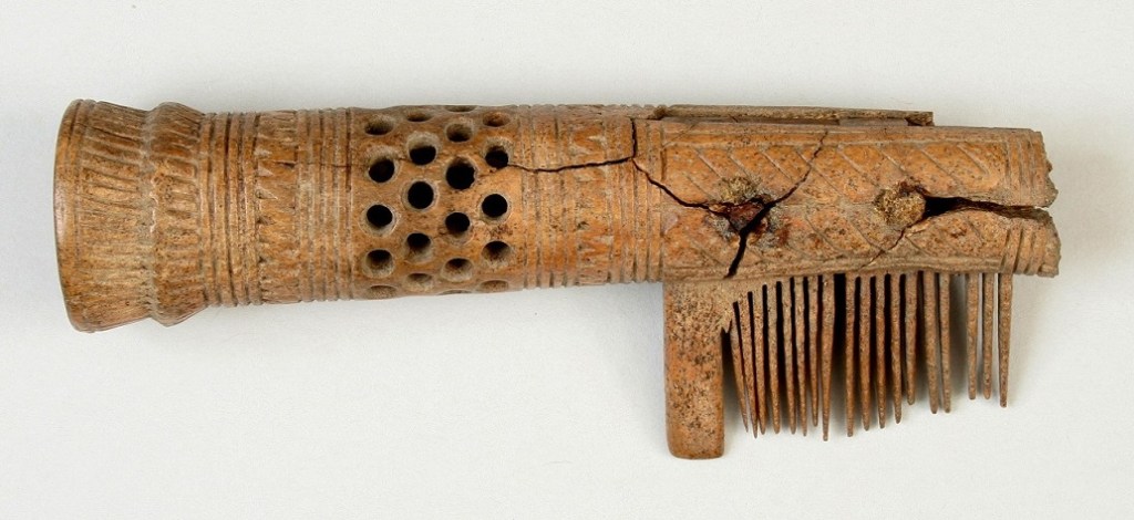 An ancient wooden comb with a carved handle, featuring decorative patterns and a series of fine teeth. The comb shows signs of aging and cracking.