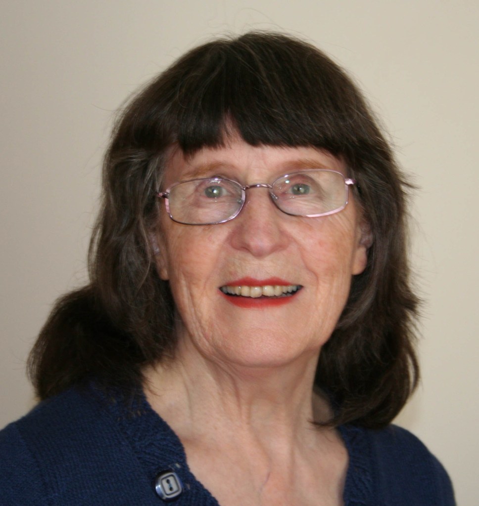 A smiling elderly woman with glasses and long dark hair, wearing a blue cardigan, against a plain background.